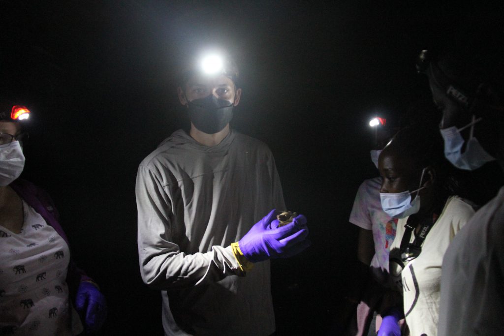 A student holding a bat at night in Africa while wearing a face mask and a headlamp. Other students look on.
